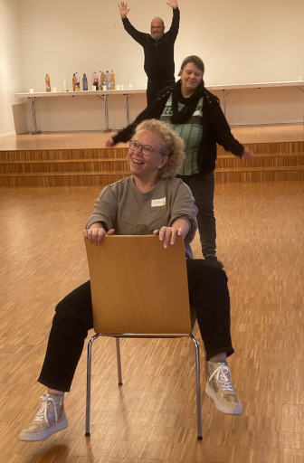 Esther sitzt auf Stuhl und macht Radelbewegung. Im Hintergrund Béat als Baum und Elke als Fussgängerin